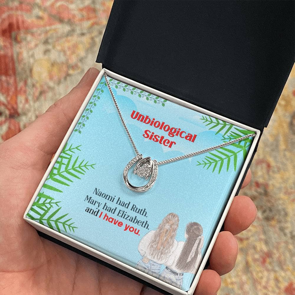 A hand holding a Lucky In Love Custom Unbiological Sisters Necklace in a box with LED lighting. The necklace features an interlocking heart pendant symbolizing the bond between unbiological sisters.