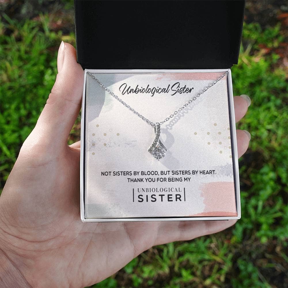 A hand holding a Customizable Unbiological Sisters Love Knot Necklace in a box, featuring a radiant cubic zirconia cushion-cut pendant.