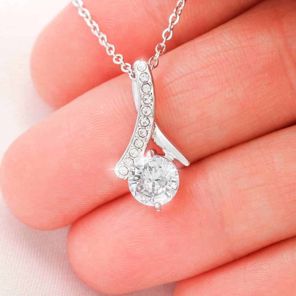 A hand holding a Customizable Unbiological Sisters Love Knot Necklace, featuring a radiant cubic zirconia cushion-cut pendant in 14k white gold or 18k gold finish. A symbol of sisterhood and love, this necklace is elegantly packaged in a mahogany-style box with LED lighting.