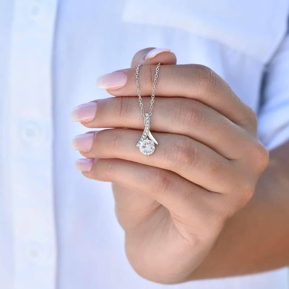 A hand holding a Customizable Unbiological Sisters Love Knot Necklace, featuring a radiant cubic zirconia cushion-cut pendant in 14k white gold or 18k gold finish. Symbolizing sisterhood and love, this necklace is a heartfelt keepsake for cherished moments.