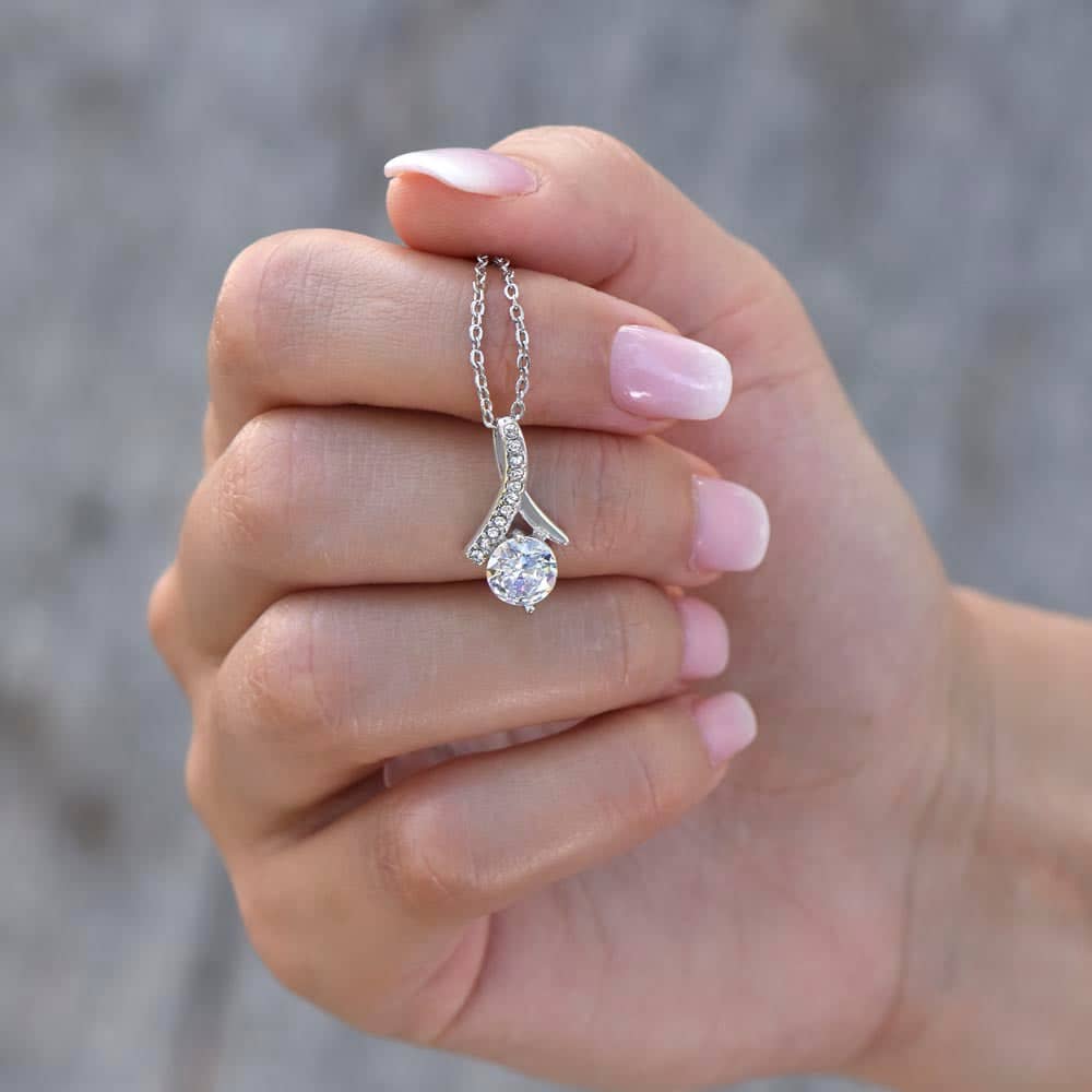 A hand holding a Customizable Unbiological Sisters Love Knot Necklace, featuring a radiant cubic zirconia cushion-cut pendant in 14k white gold or 18k gold finish. A symbol of sisterhood and love.