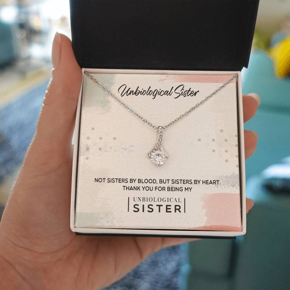 A hand holding a Customizable Unbiological Sisters Love Knot Necklace in a box, featuring a radiant cubic zirconia cushion-cut pendant.