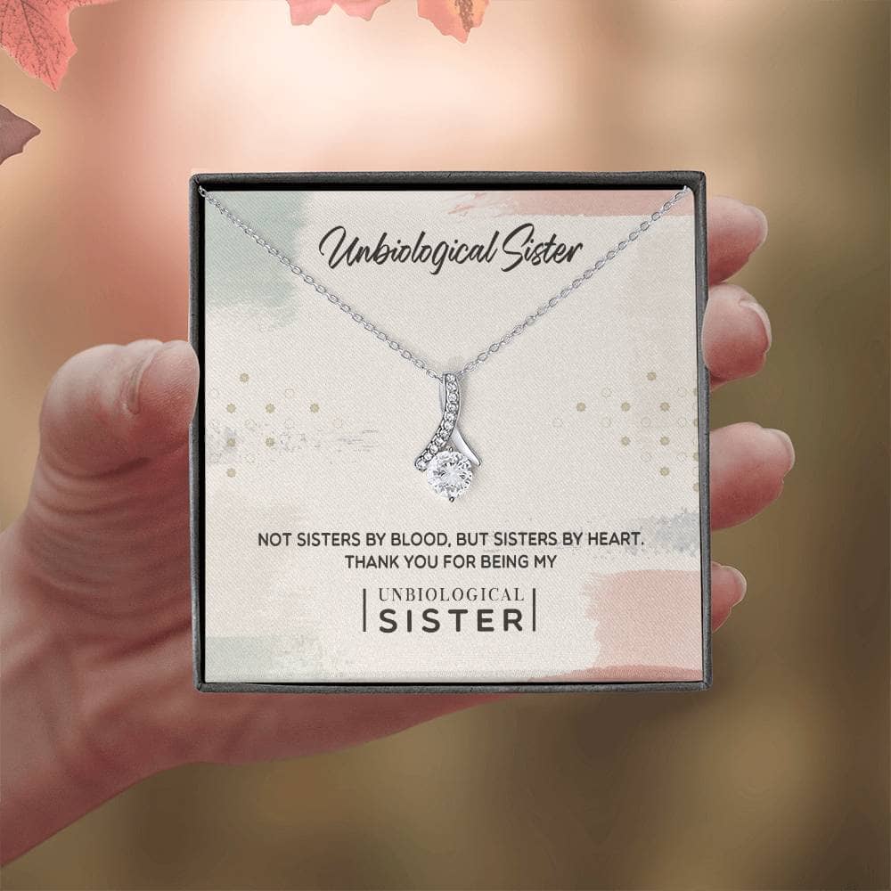 A hand holding a Customizable Unbiological Sisters Love Knot Necklace, featuring a radiant cubic zirconia cushion-cut on a heart or knot pendant in 14k white gold or 18k gold finish.