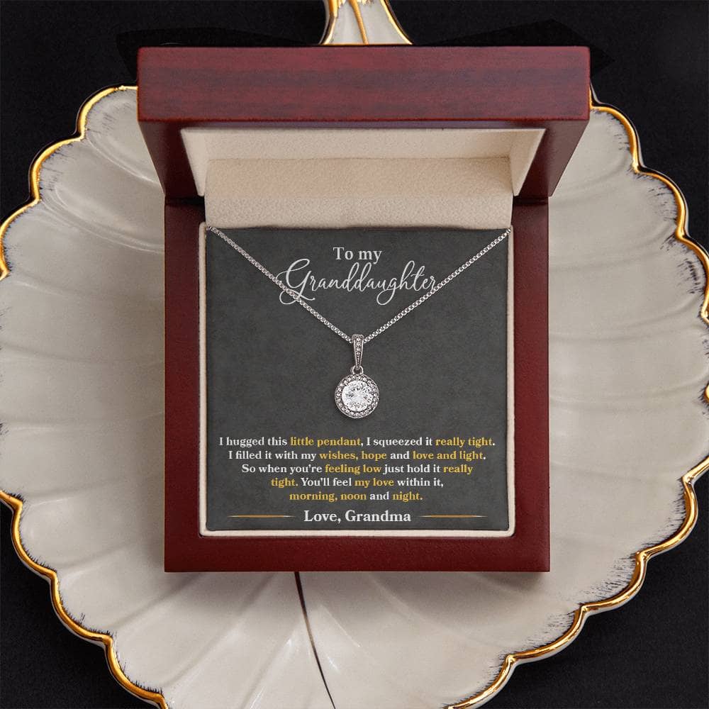 A necklace in a box on a plate, featuring the Personalized Granddaughter Heart Necklace. A timeless and elegant gift with a cushion-cut center cubic zirconia that sparkles with every step. Crafted with a 14k white gold finish over stainless steel, adorned with brilliant CZ crystals. Adjustable box chain length of 16" - 18". Lovingly packaged in a soft touch box for easy gifting.