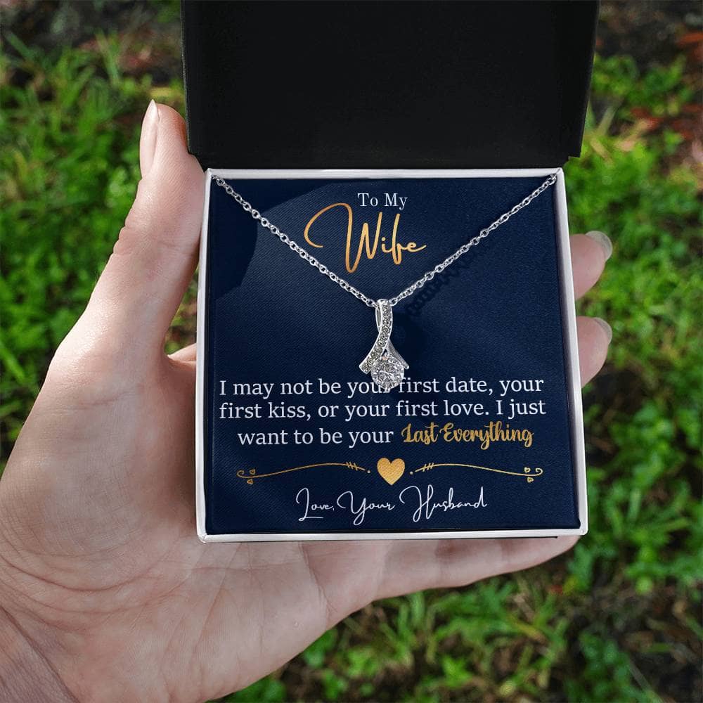 Alt text: "Personalized Wife Necklace: A hand holding a ribbon-shaped pendant in a box, adorned with a 7mm cubic zirconia. Symbol of love and connection."