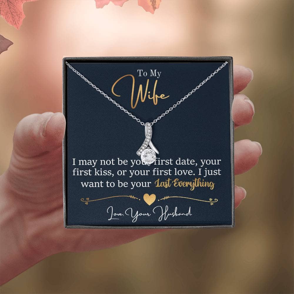 A hand holding the Personalized Wife Necklace: Symbol Of Love And Connection, a ribbon-shaped pendant crafted in 14k white gold or 18k yellow gold over stainless steel. Adorned with a 7mm cubic zirconia, it exudes sophistication and grace. Adjustable chain length of 18" - 22".